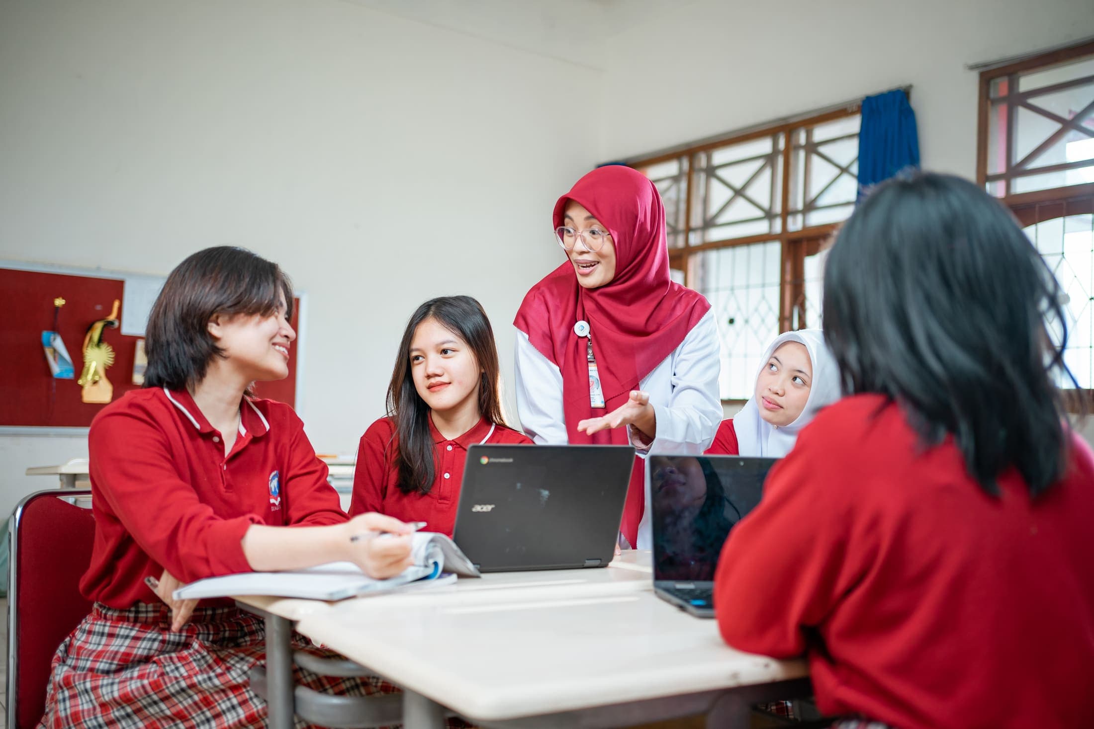 A teacher guiding a group of students working with laptops in a bright classroom